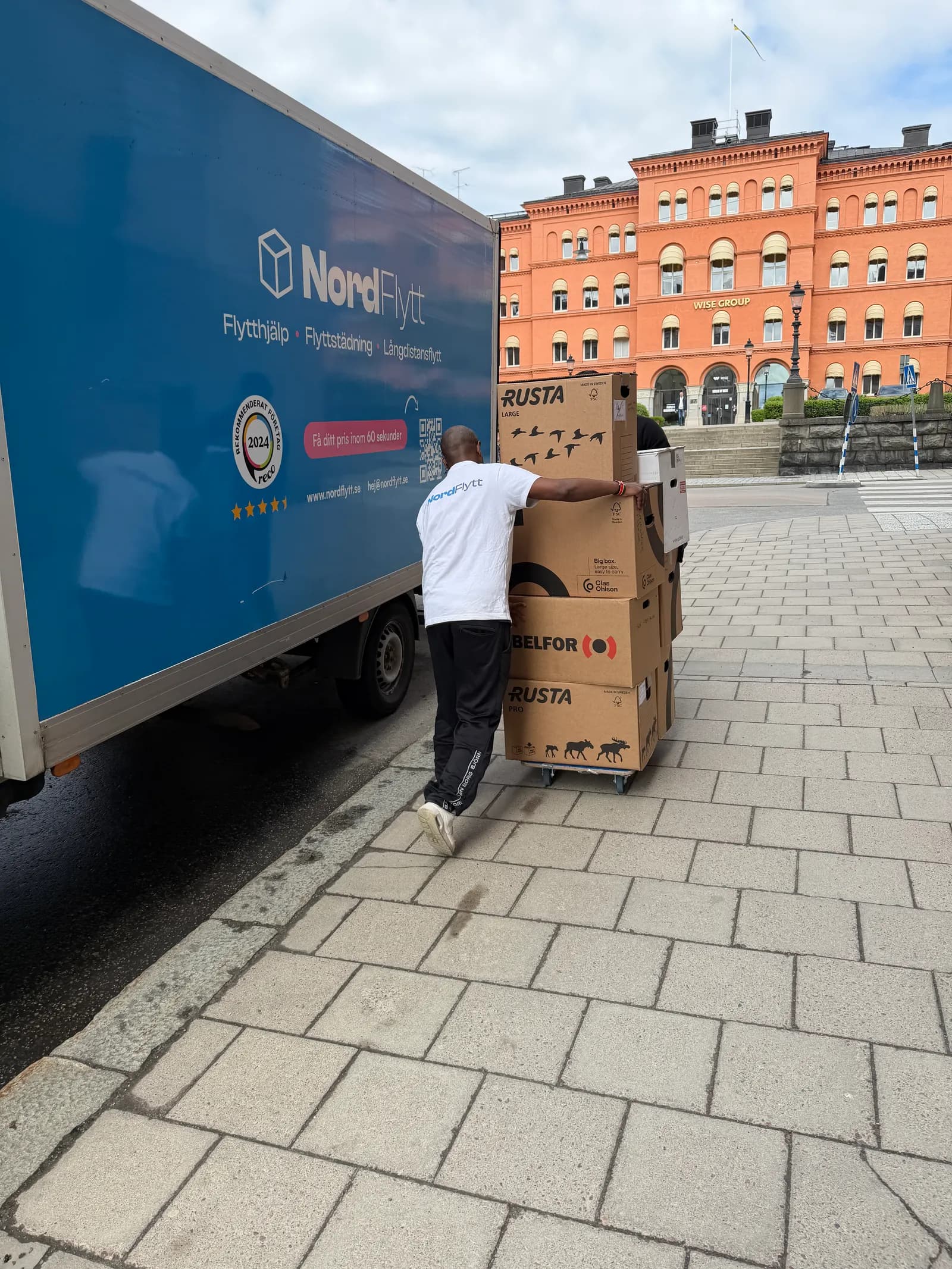 Flyttare från Nordflytt bär en flyttkartong vid en blå Nordflytt-lastbil på en gata i Stockholm.