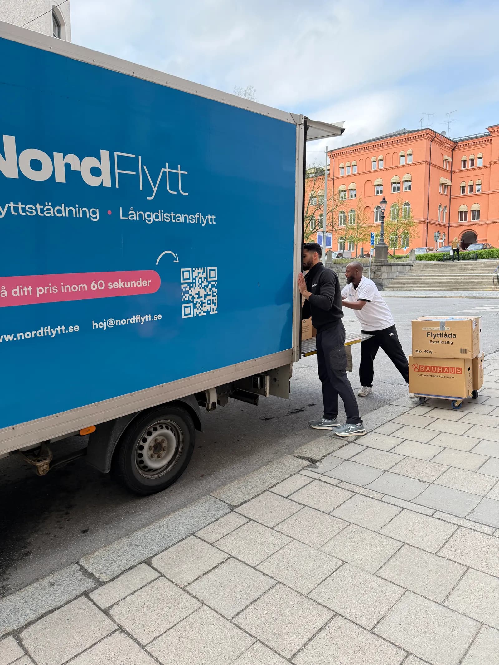 Nordflytts blå flyttbil med tjänstetext och flyttare på en gata i Stockholm.